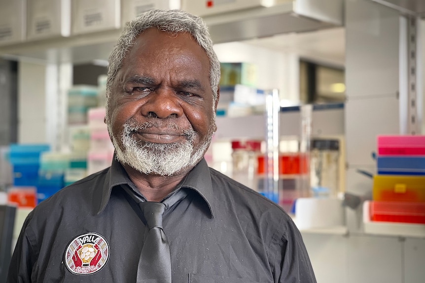 Ross wearing a black shirt with a "YALU Aboriginal Corporation" logo, standing in a laboratory setting. 