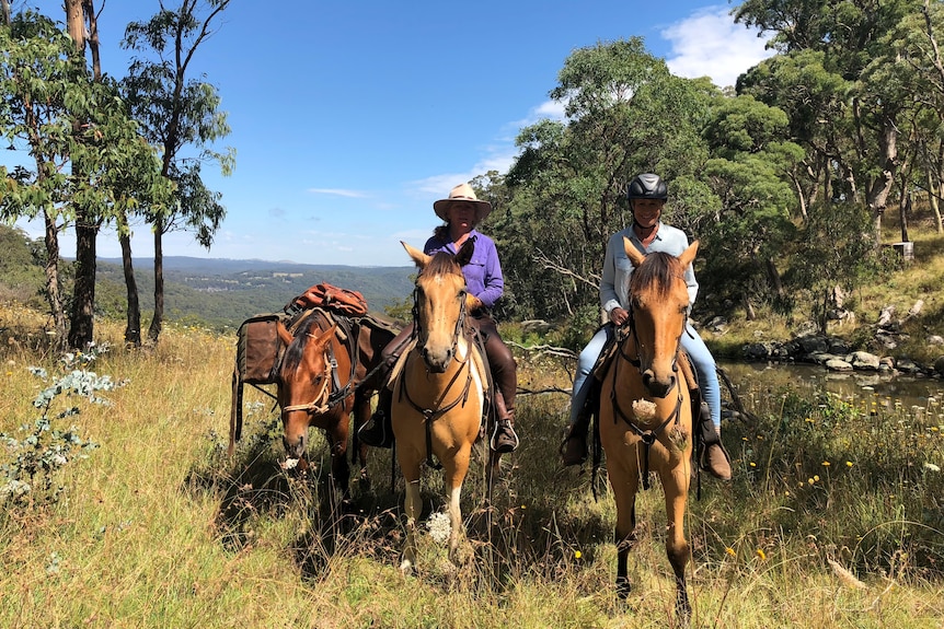 Two women riding horses in the countryside, large open vista of valley below in the background. 