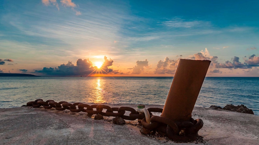 The sun sets over the ocean, with an anchor and large chain on a jetty in the foreground.