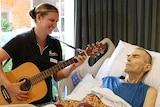 A woman holds a guitar by the bedside of a sick man