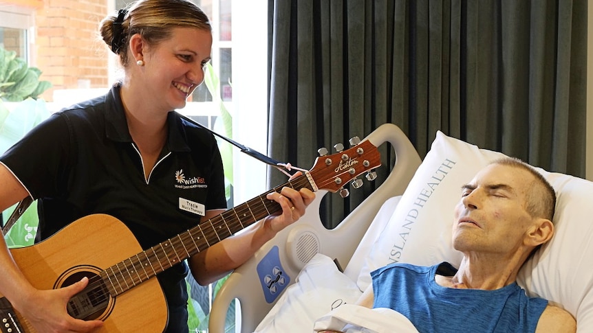 A woman holds a guitar by the bedside of a sick man