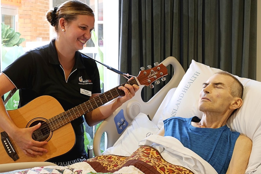 A woman holds a guitar by the bedside of a sick man