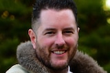 Marcus Stewart smiles as he stands outside, wearing a shirt, tie and possum-skin cloak.