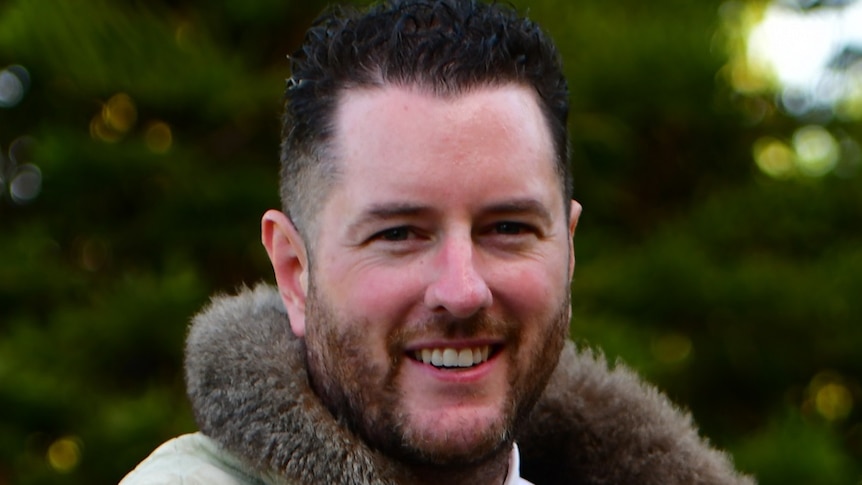 Marcus Stewart smiles as he stands outside, wearing a shirt, tie and possum-skin cloak.