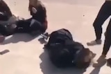 One girl lies on the ground while another kicks her head and others sit on the concrete ground