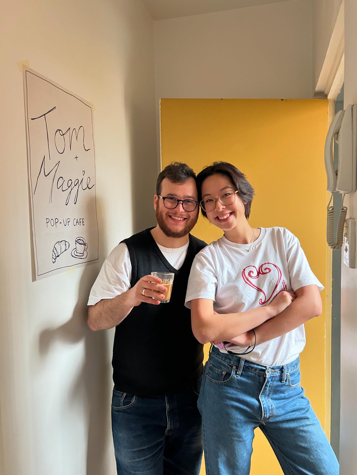A picture of young couple posing by a yellow apartment door, next to a sign reading 'Tom plus Maggie's pop-up cafe'.