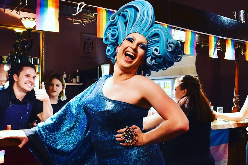A drag queen wearing a blue dress with mardi gras flags in the background 