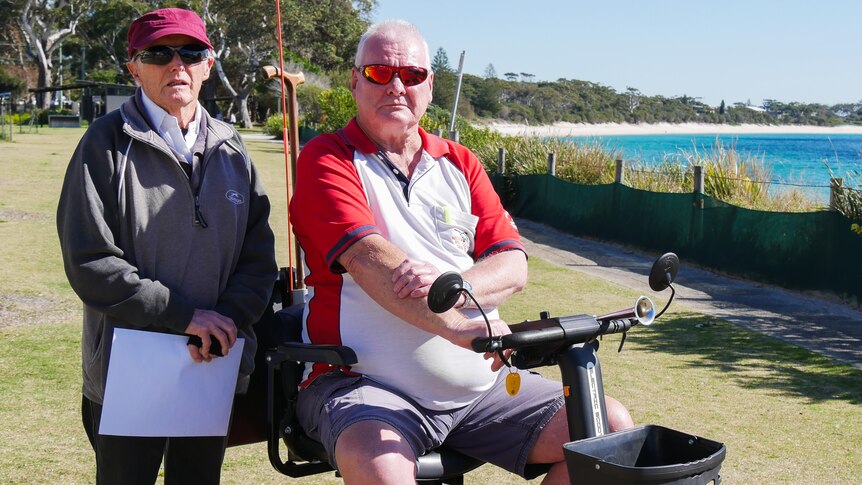 An elderly man standing next to another elderly man who is sitting on a mobility scooter next to a beach.