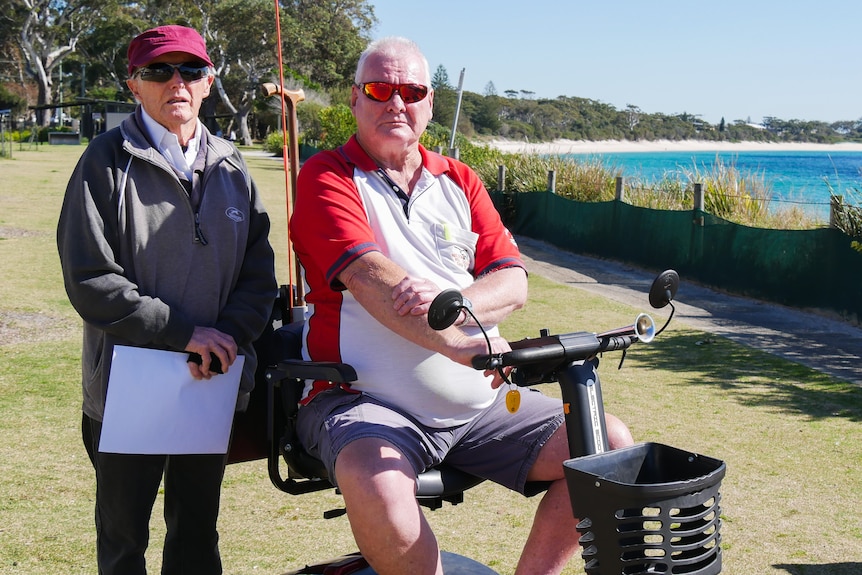 An elderly man standing next to another elderly man who is sitting on a mobility scooter next to a beach.