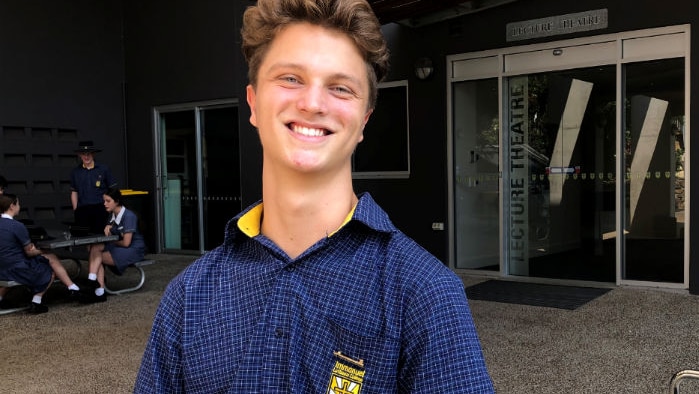 High school student standing in front of library with other students at table in background.