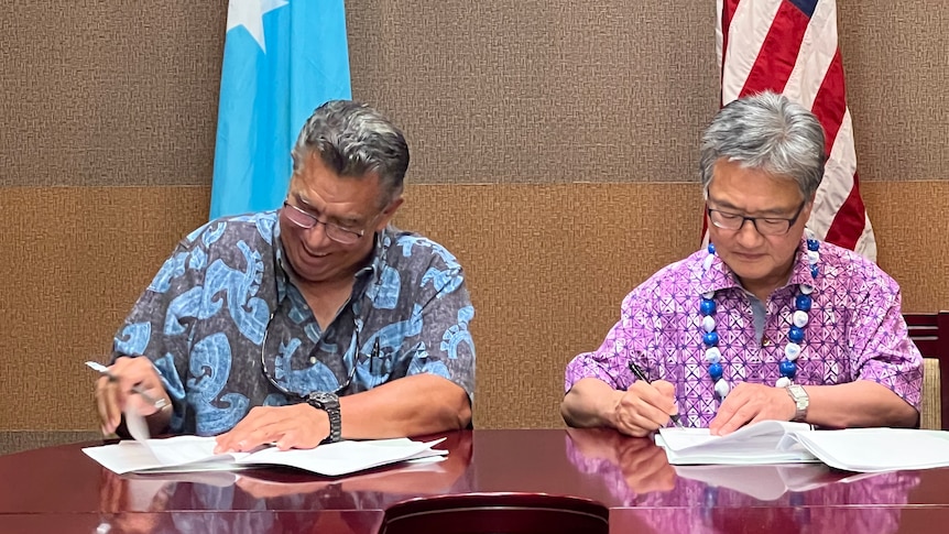 Two men in bright blue and pink shirts sign documents at desk in front of US and FSM flags.