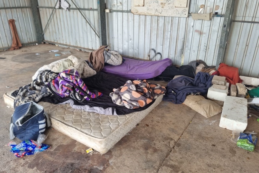 A mattress and used purple, blue and black bedding are piled on it and on the concrete floor in a shed.