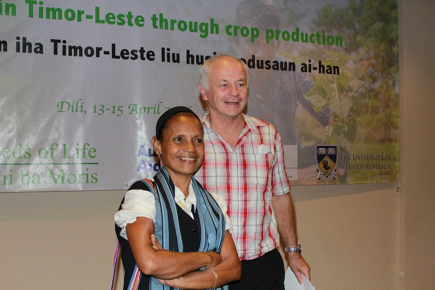 East Timorese woman in colourful weaving and black vest with an Australian man stand in front of poster, Dili East Timor