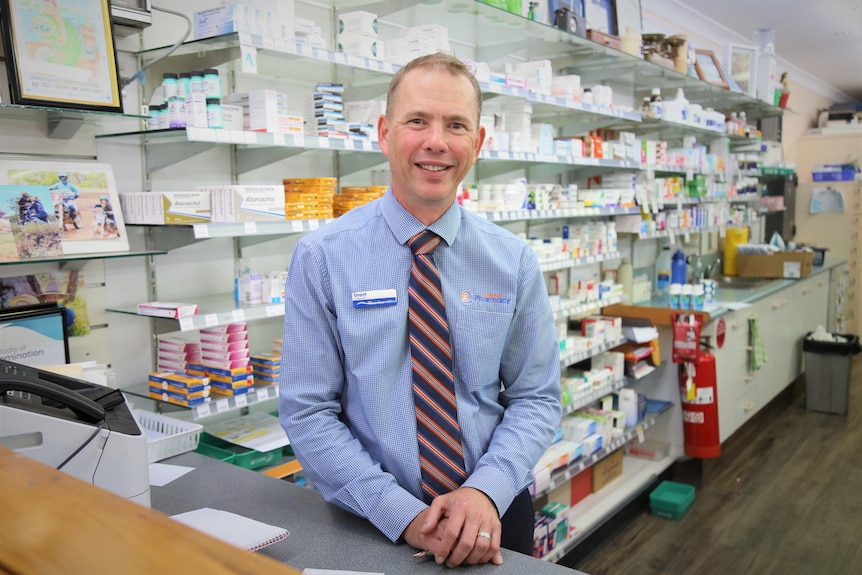 A male pharmacist stands in front of shelves of medicine boxes
