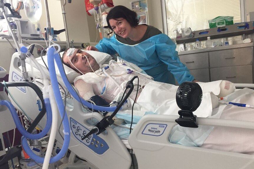 A woman leans over and smiles next to her husband who is in a hospital bed with all limbs amputated and medical cords surround