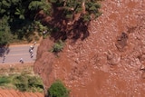 An aerial view shows brown sludge blocking a road and inundating green forest area.