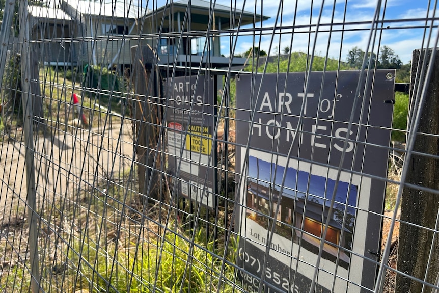 A fenced off display home with weeds on the site and a sign reading art of homes