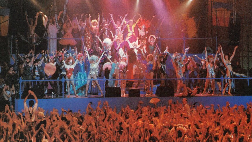 dozens of drag queens dance on a stage in front of a large crowd.