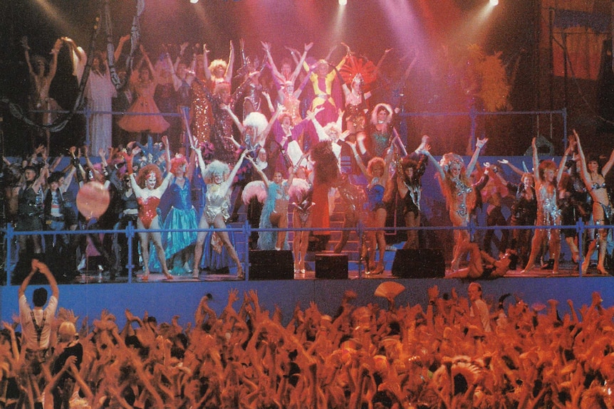 dozens of drag queens dance on a stage in front of a large crowd.