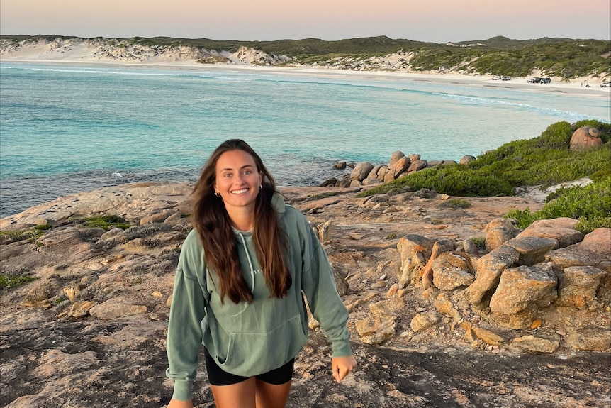 girl with long brown hair in green jumper with a beach in the background 