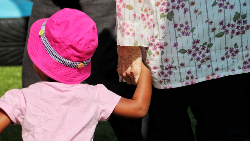 A rear photo of an elderly woman holding the hand of a small child wearing a pink hat.