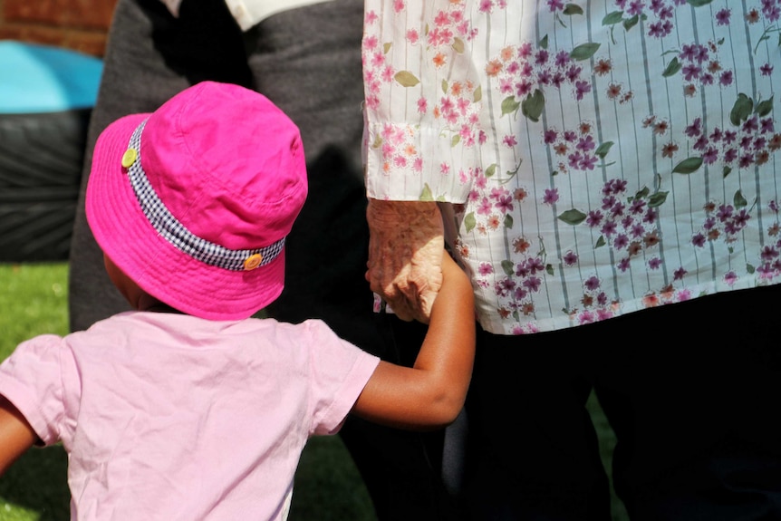 A rear photo of an elderly woman holding the hand of a small child wearing a pink hat.