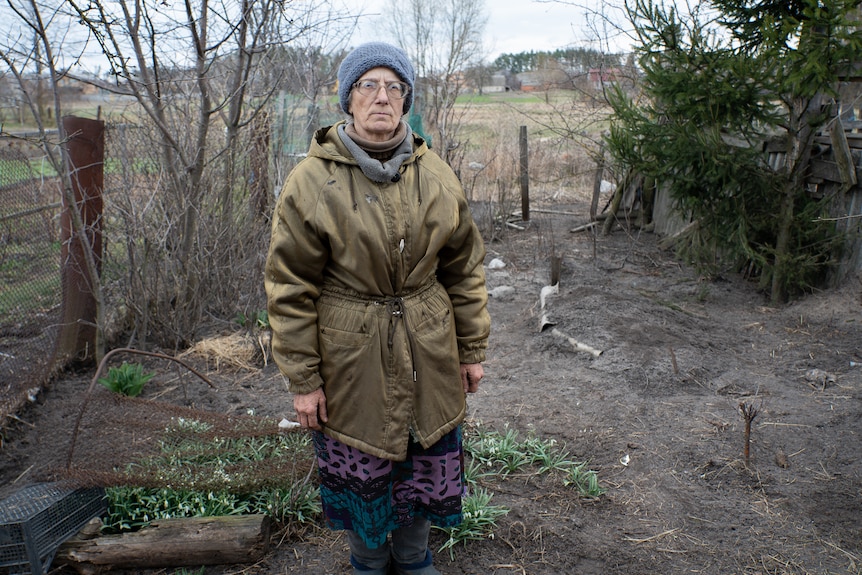 A woman solemnly poses for the camera in front of an unmarked grave in her backyard.