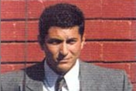 A man in a suit and tie stands in front of a red brick wall.