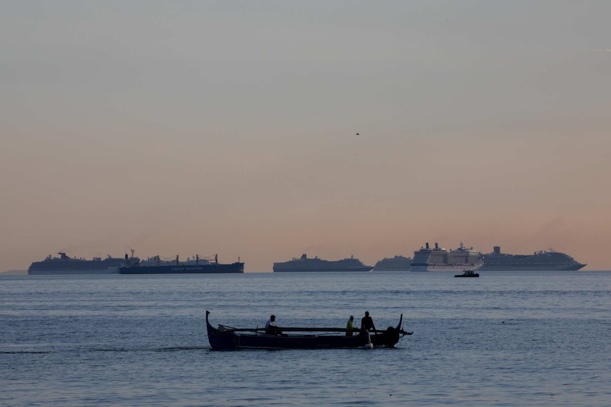 Fishermen sail past a group of cruise ships sitting on the horizon at dusk.
