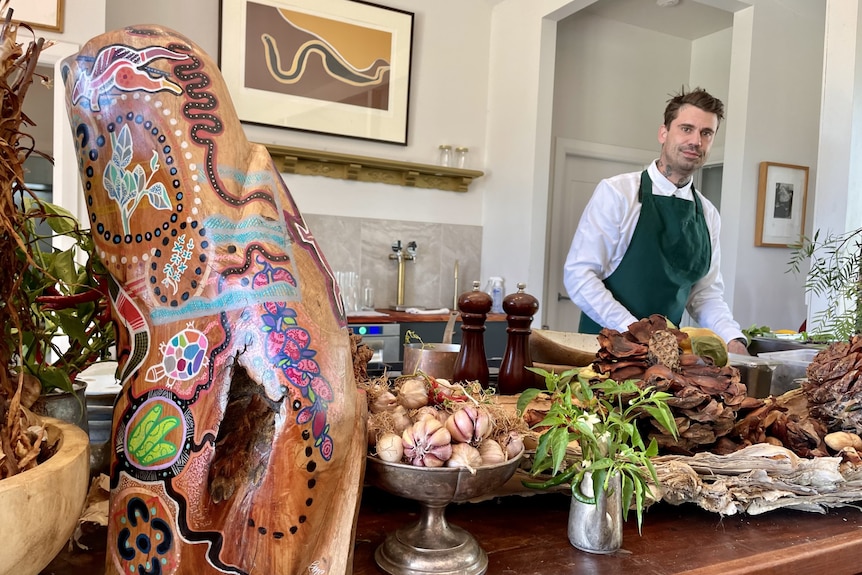 A man in a chef's apron stands in a kitchen with fresh produce surrounding him.