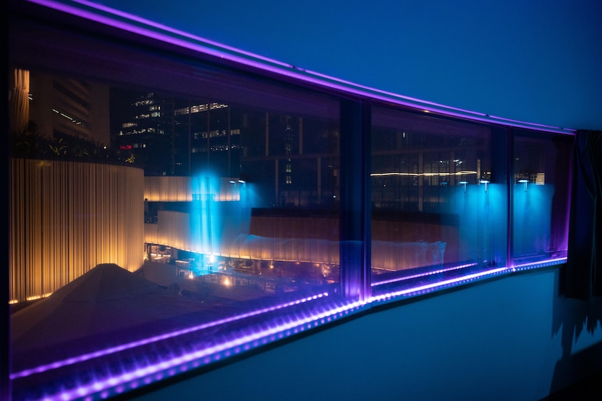 The inside of a blue neon-lit hotel room is reflected through paneled glass windows overlooking a city nightscape.