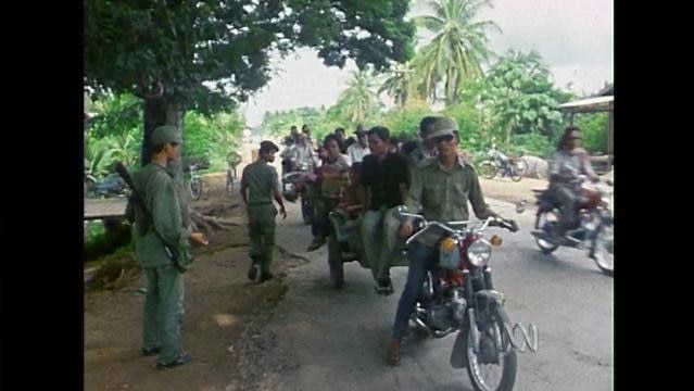 Soldiers stand beside road in Cambodia as people on motorbikes