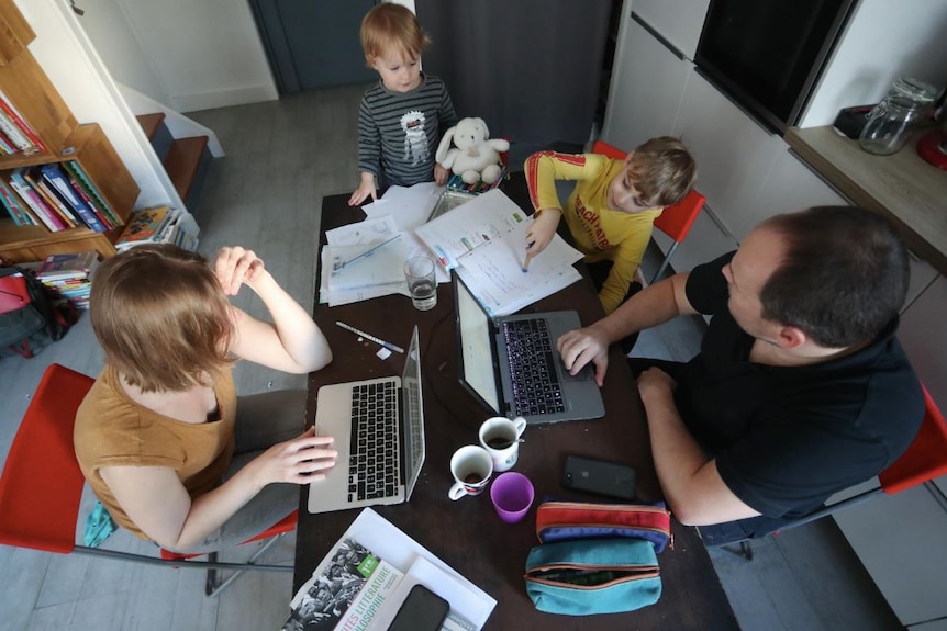 A view from above shows two parents working at the dining table, surrounded by papers, pencil cases and their young children.