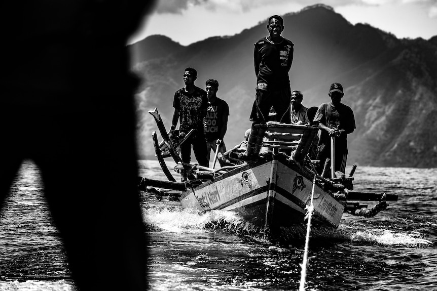 Five men stand on a boat at sea with mountains in the background.