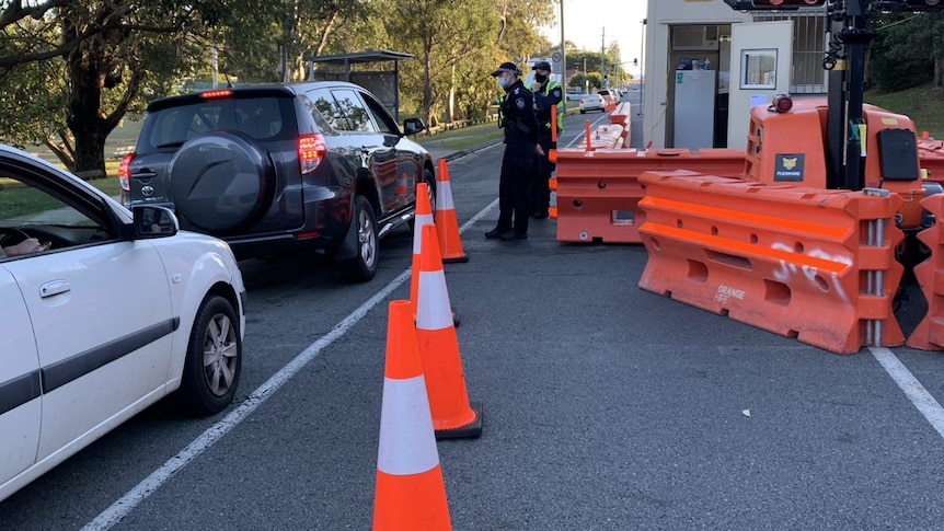 Cars lined up being checked by police in black wearing masks, orange witches hats and barriers on road