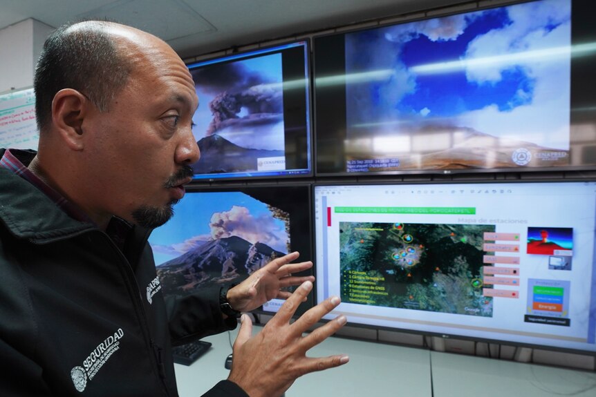 A man gestures at a row of monitors showing a volcano spewing ash, and seismological readings.