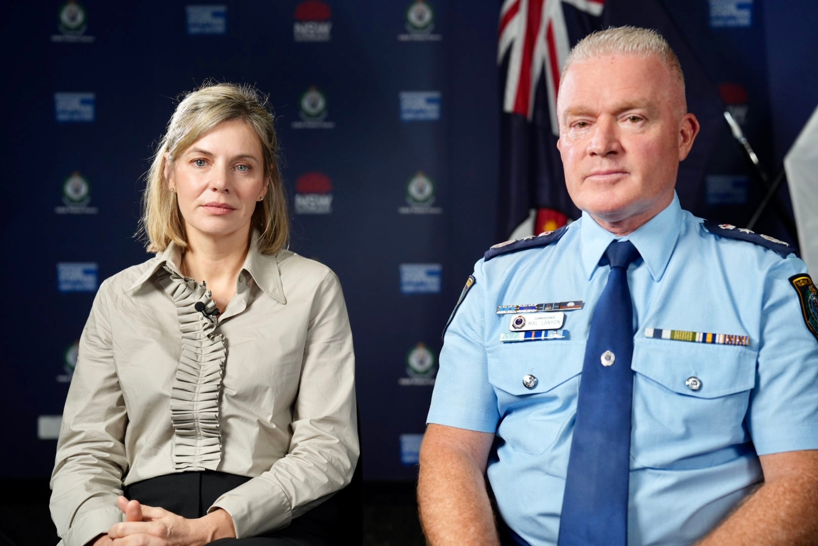 Mal Lanyon to the right in his blue police uniform, and Kristen Hilton in a beige long sleeve top. Both are serious, sitting.