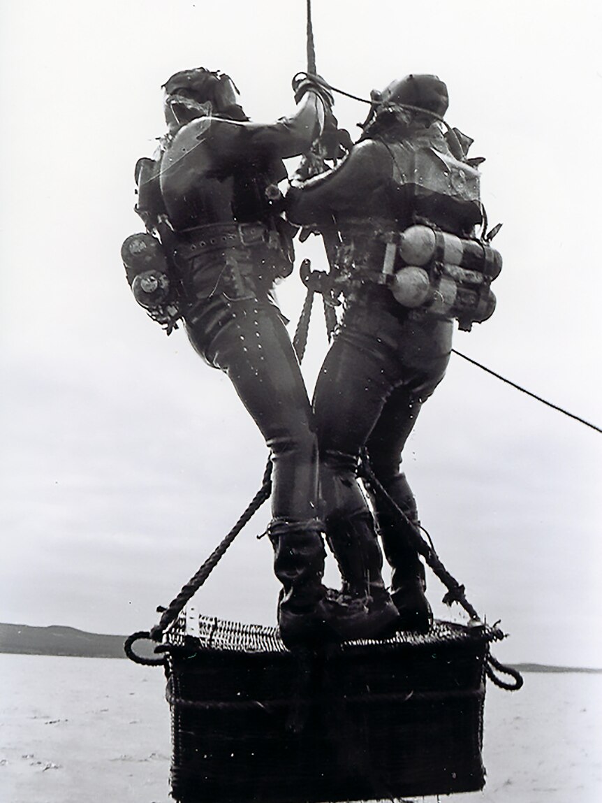 Two divers wearing heavy lead weights to allow them to work in heavy tides stand on a basket being winched up out of the water