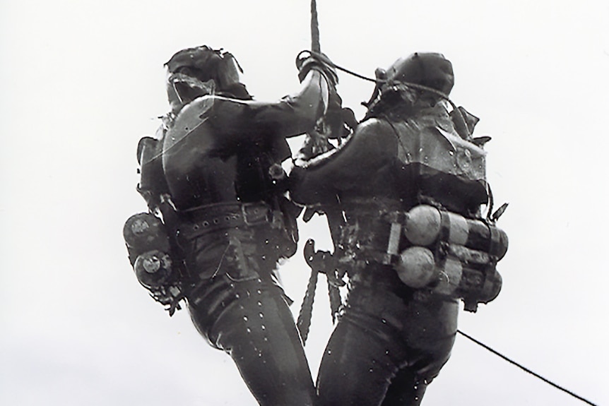 Two old-style divers standing on a square basket being winched up in the air