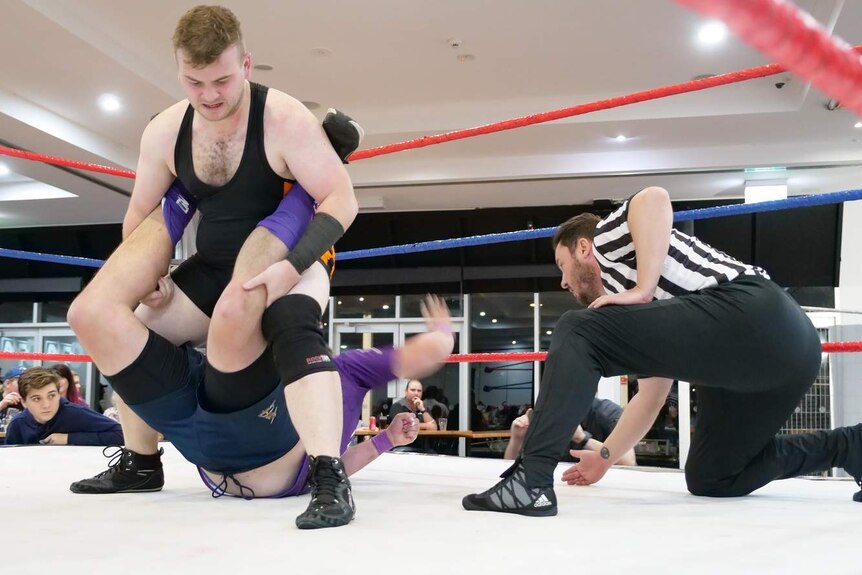 A wrestling referee kneels in a wrestling ring while two men wrestle.