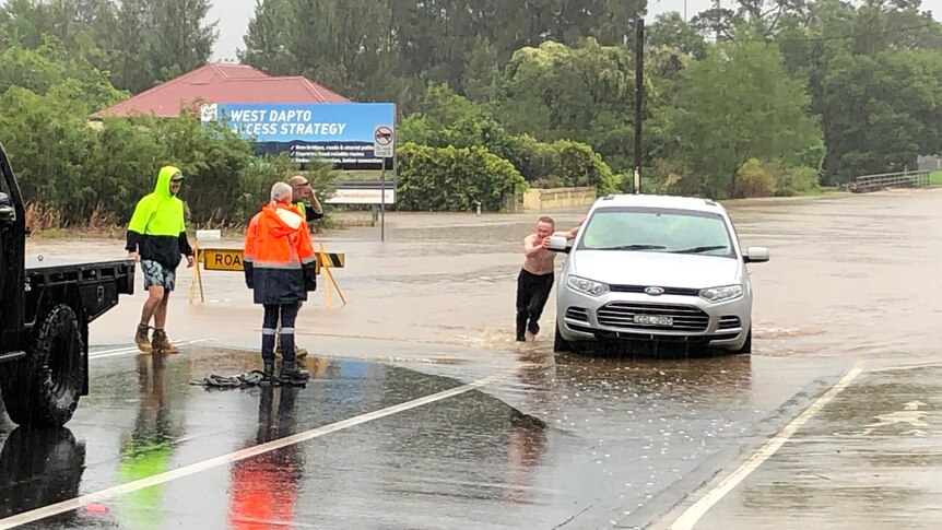 A car being pushed by a shirtless man through floodwater.