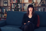 A woman sits on a couch with a bookshelf behind her.