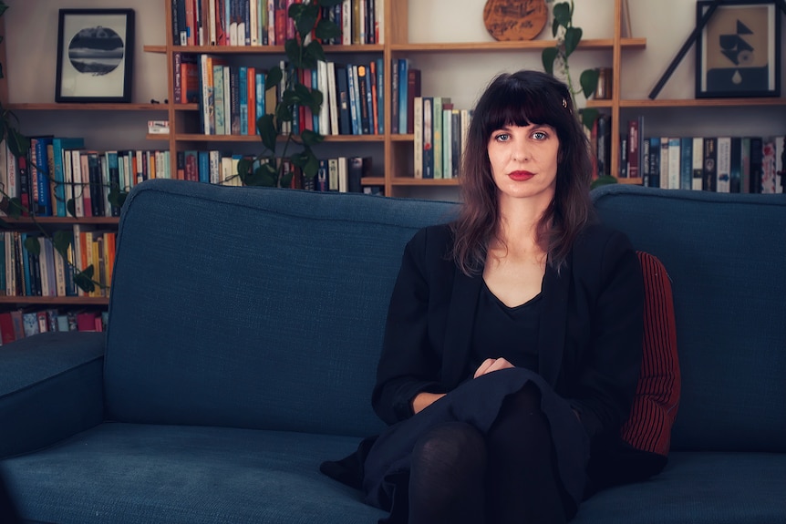 A woman sits on a couch with a bookshelf behind her.