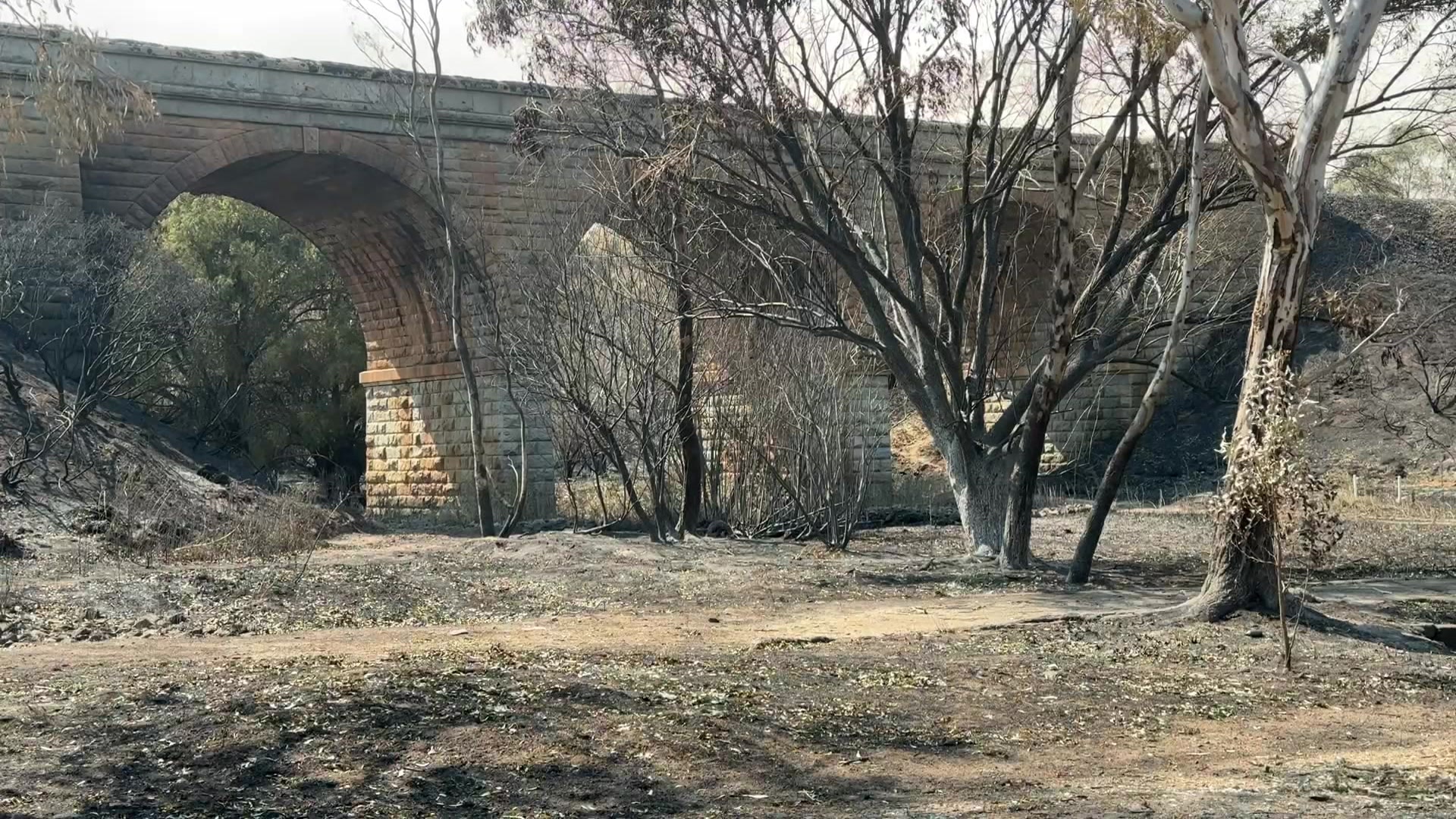 Uma ponte de pedra cercada por árvores carbonizadas e terra enegrecida.