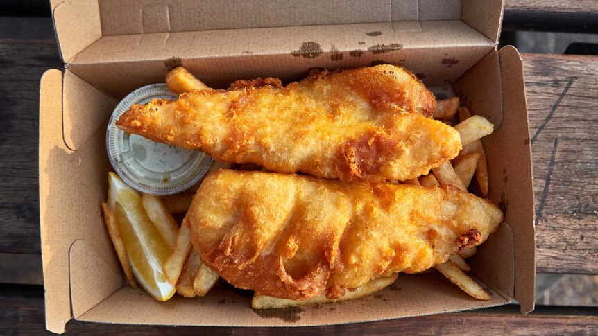 A cardboard takeaway box on a picnic table holds golden, fried fish atop chips with a slice of lemon and tartare sauce.