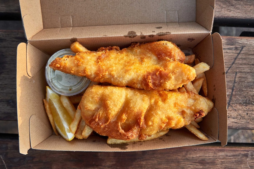 A cardboard takeaway box on a picnic table holds golden, fried fish atop chips with a slice of lemon and tartare sauce.