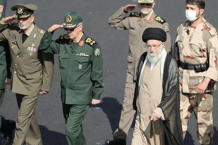 Ayatollah Ali Khamenei wearing robes uses a walking stick while walking with men dressed in military uniform.