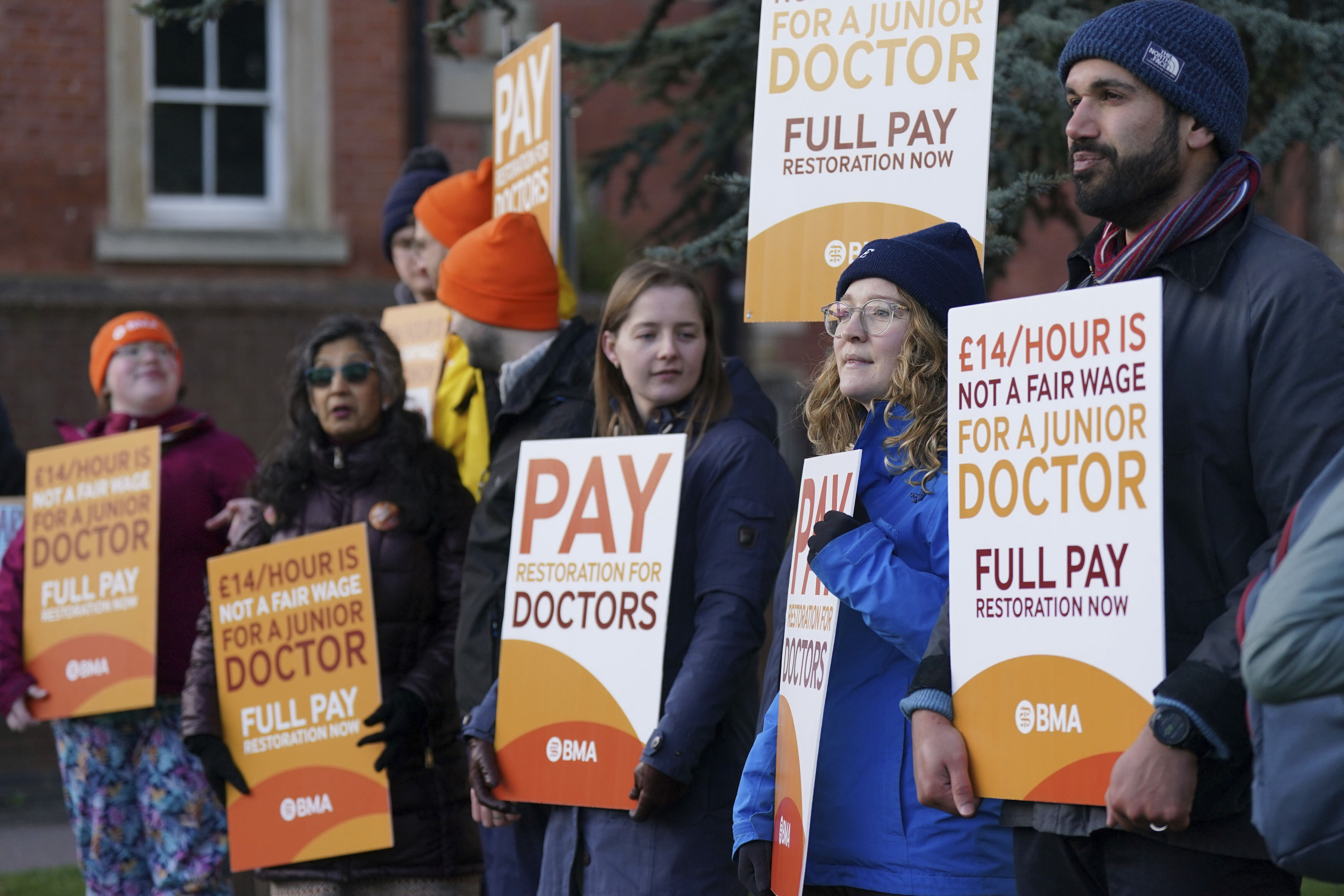 A small group of young people in winter clothing stand outside a brick building holding orange signs demanding fair wages.
