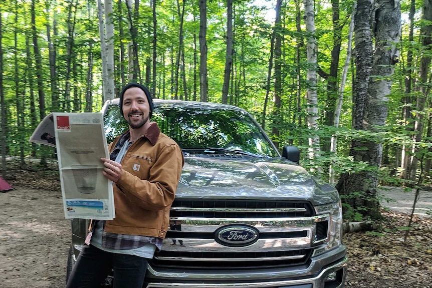 Alex McClintock smiles and stands beside a four-wheel drive, while holding a newspaper.