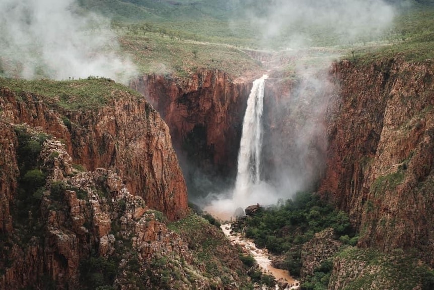 A waterfall flows into a creek near lake argyle, with spectacular red cliffs and a green landscape in the background.